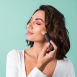 mulher jovem em um vestido de noite branco em verde claro com maquiagem rosa brilhante segurando um pincel de blush Easy Resize.com